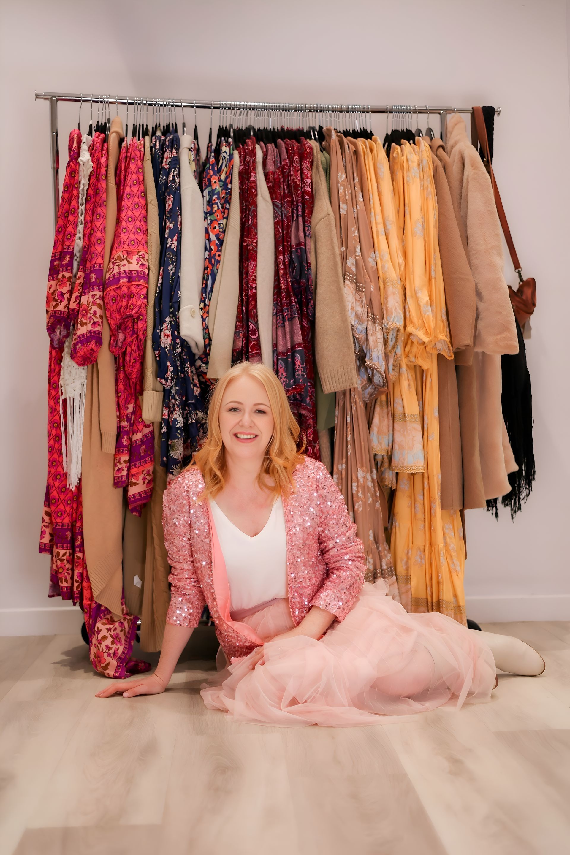 Woman in pink sparkly jacket and skirt sits in front of clothing rack with colourful garments.