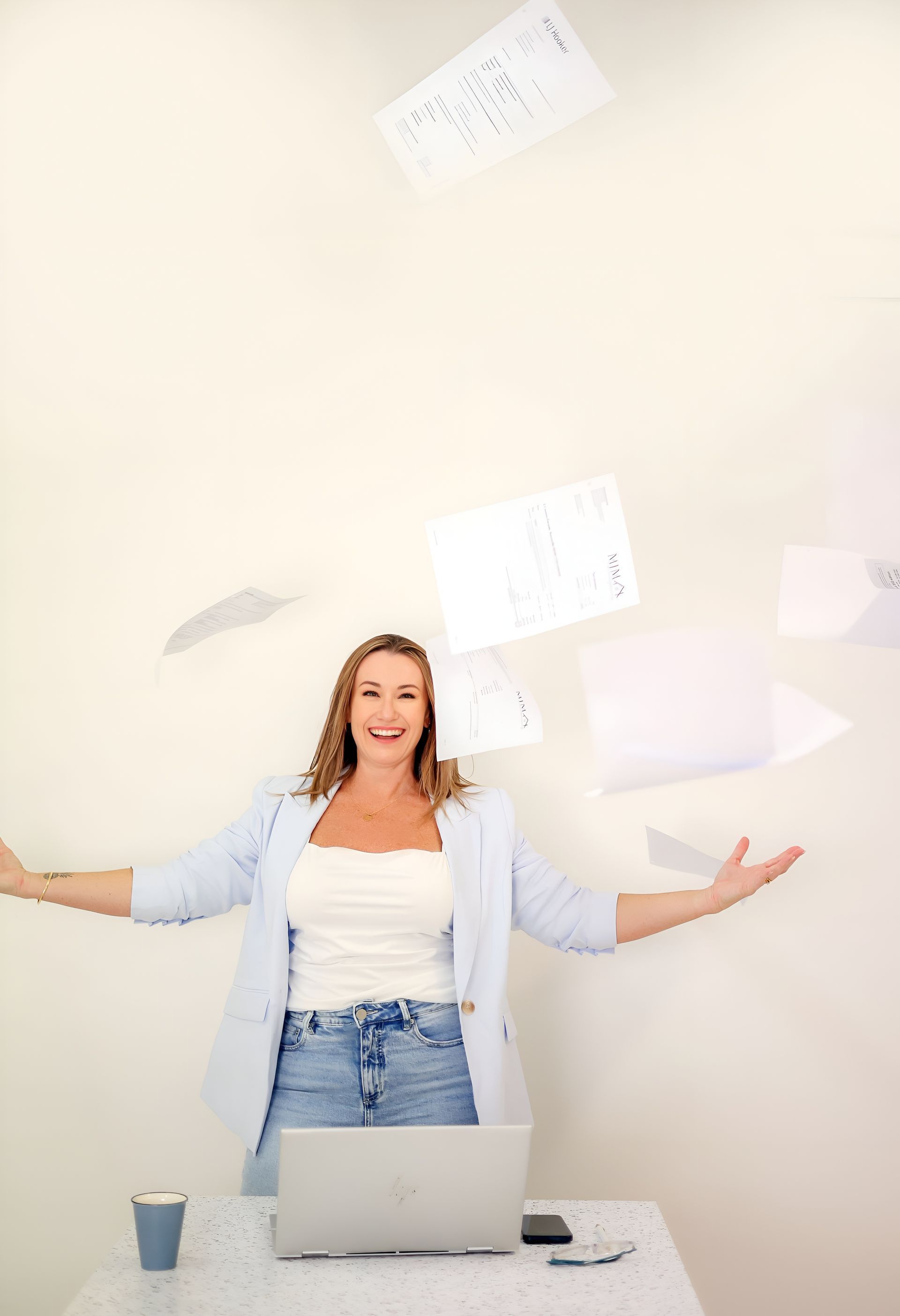 Woman tossing papers in the air, smiling, standing at a desk with a laptop and coffee. Light blue blazer.