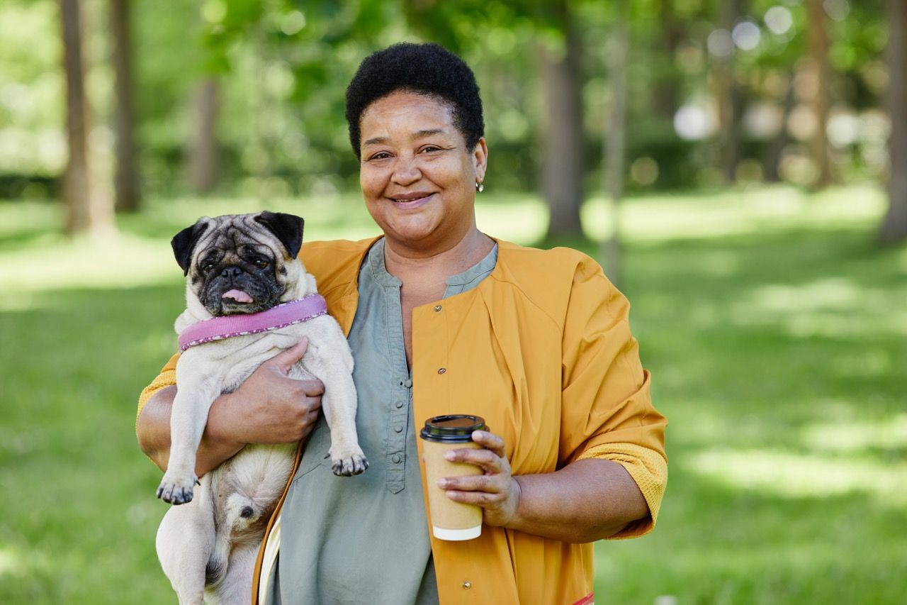 Woman holding a pug dog, smiling, with coffee, in a park.