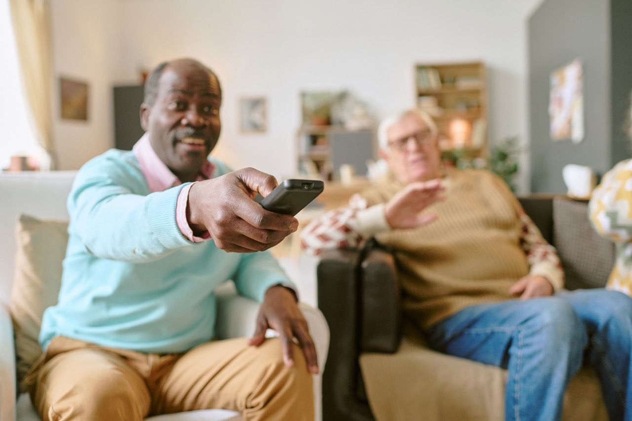 Man holding remote, pointing, smiling; another man sits nearby, gesturing, in a living room.