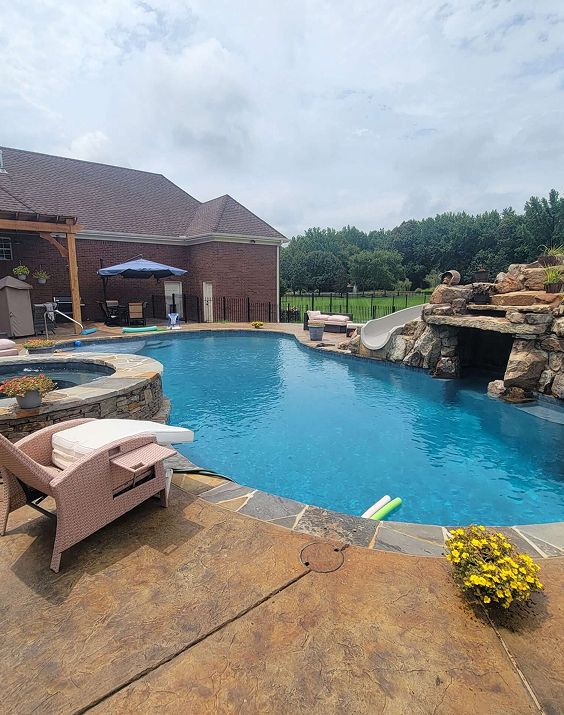 A swimming pool with a slide, waterfall, and surrounding patio in front of a brick house. Blue water, sunny day.