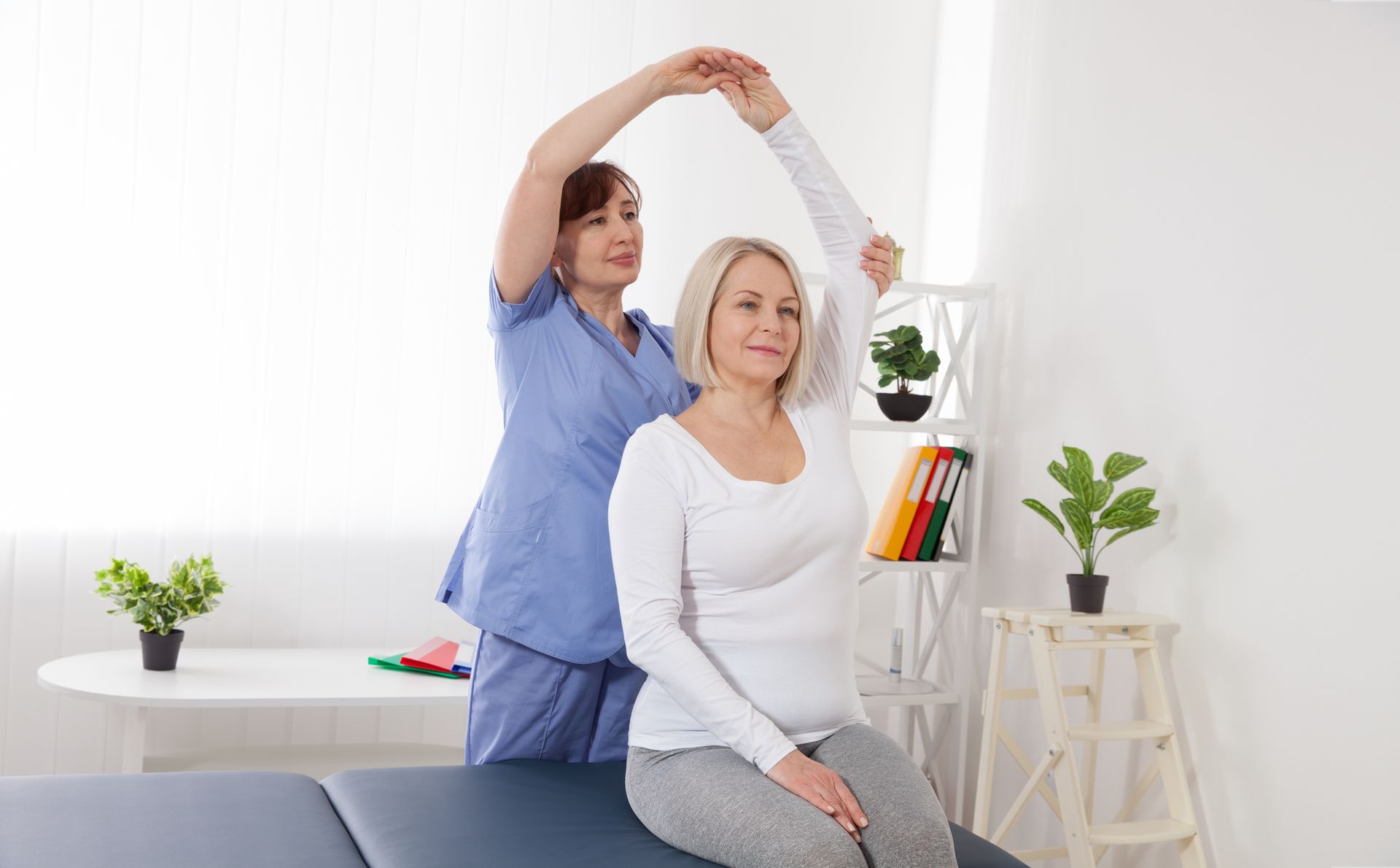 Physical therapist assisting a seated woman with an overhead stretch in a bright clinic