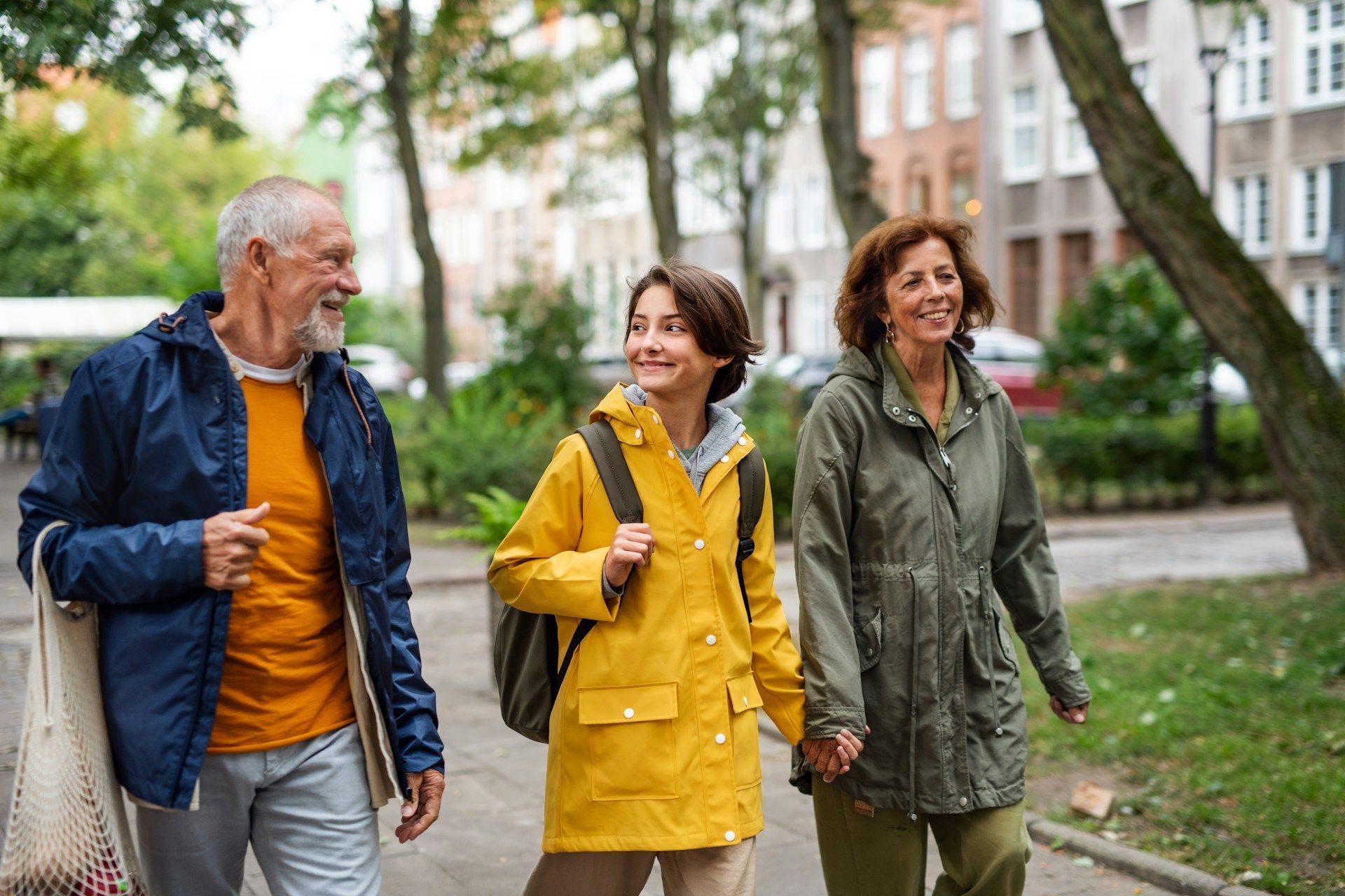 A smiling family of three walking down a sidewalk. The child in yellow raincoat and carrying a backpack.