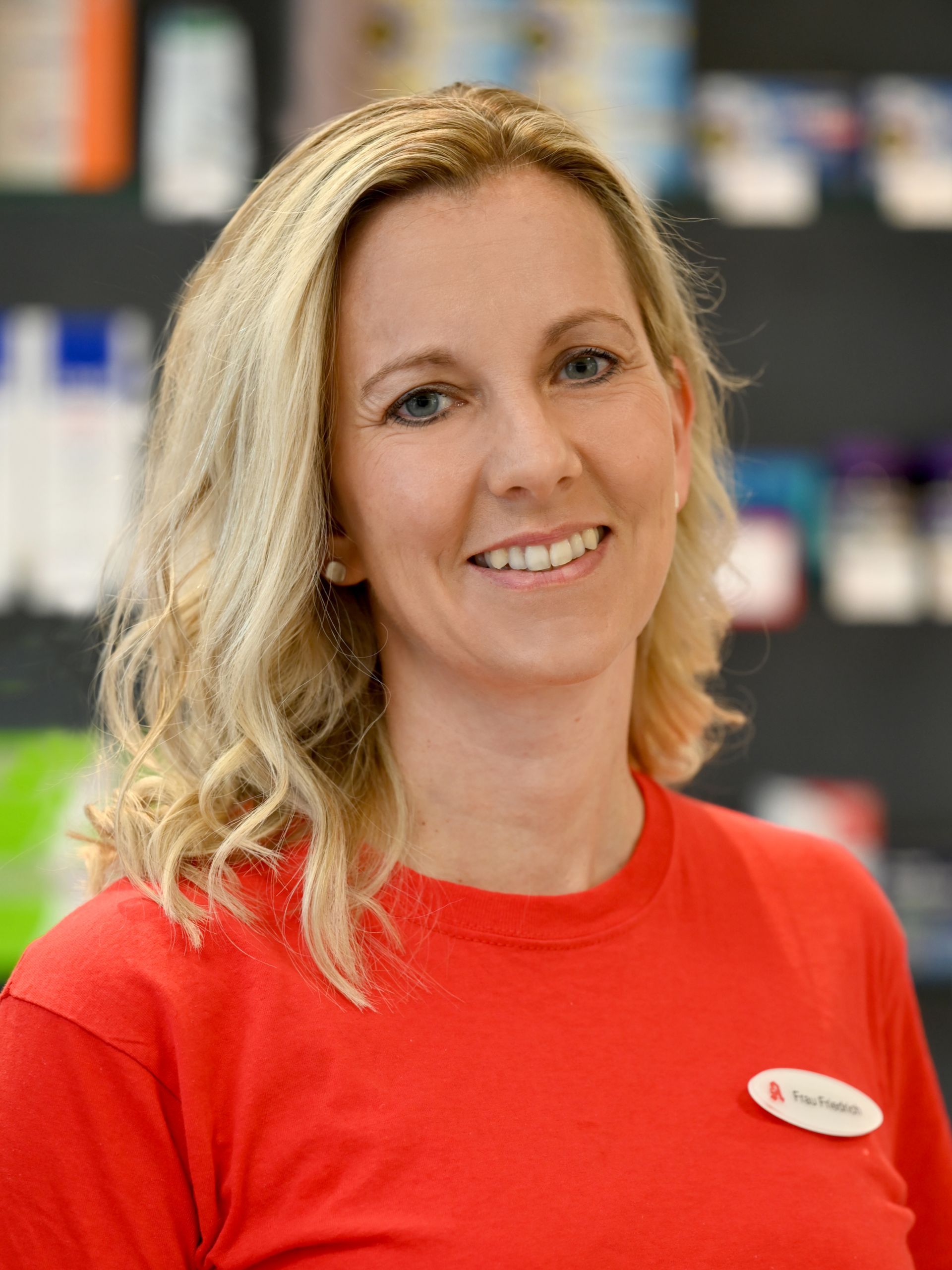 Blonde Frau mit einem leichten Lächeln in einem roten Hemd, wahrscheinlich eine Apothekerin, in einer Apotheke.
