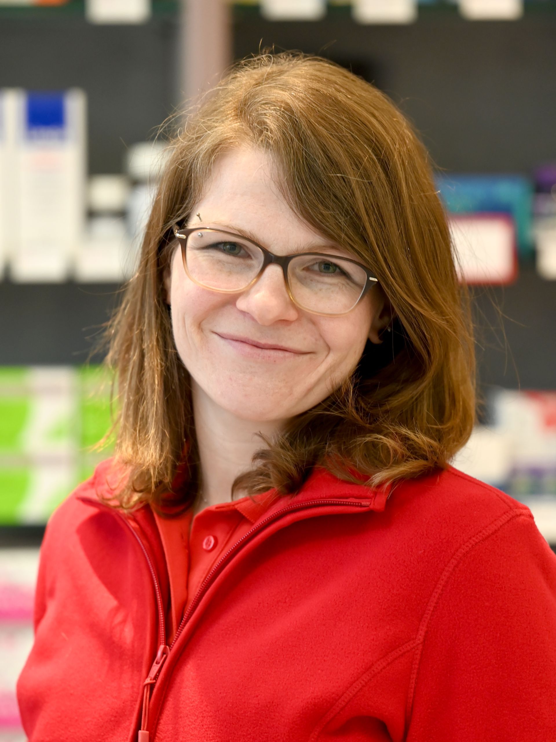 Frau mit Brille und rotem Pullover lächelt in einer Apotheke.