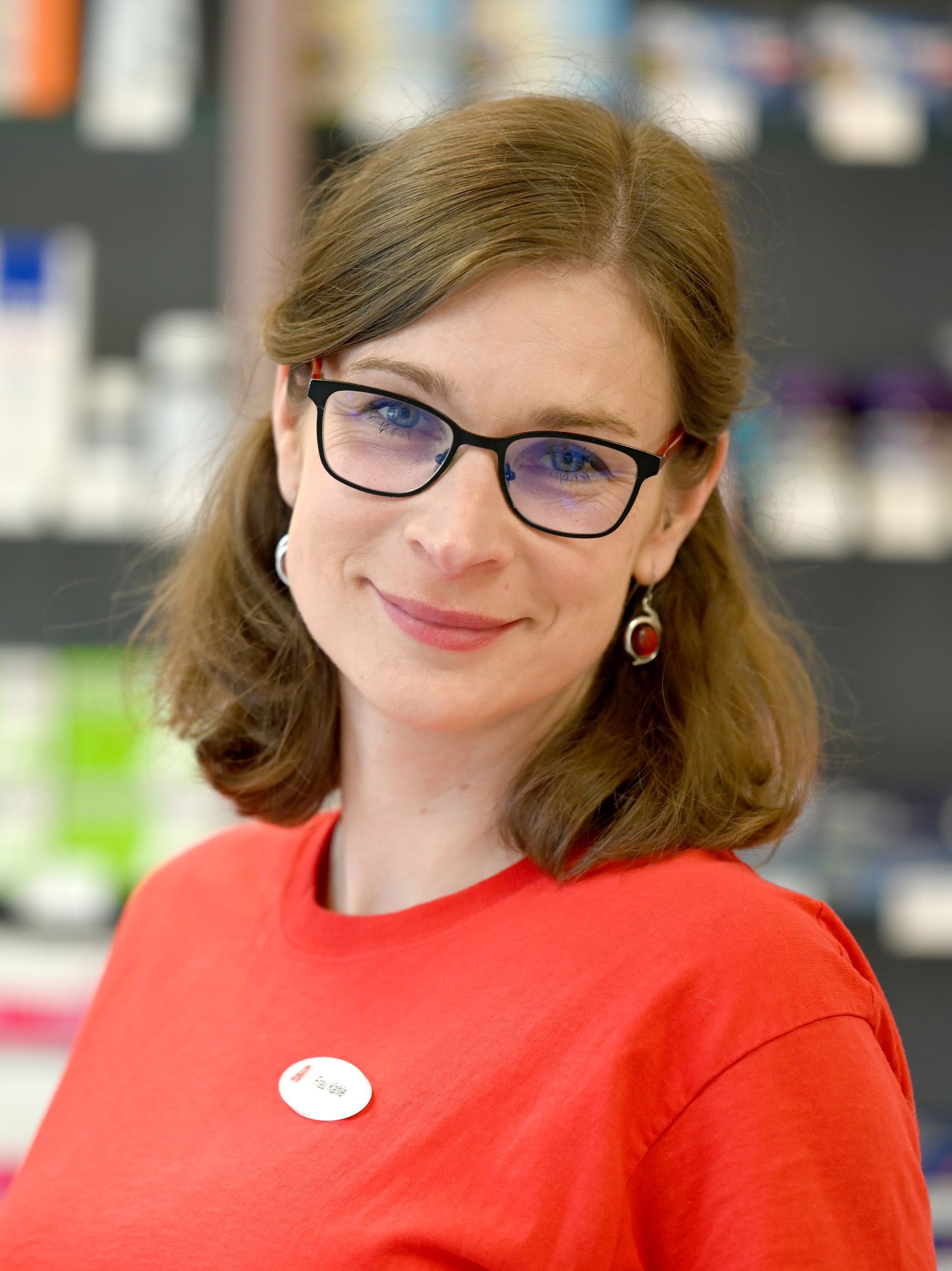 Frau mit Brille, rotem Hemd, lächelnd in einer Apotheke.