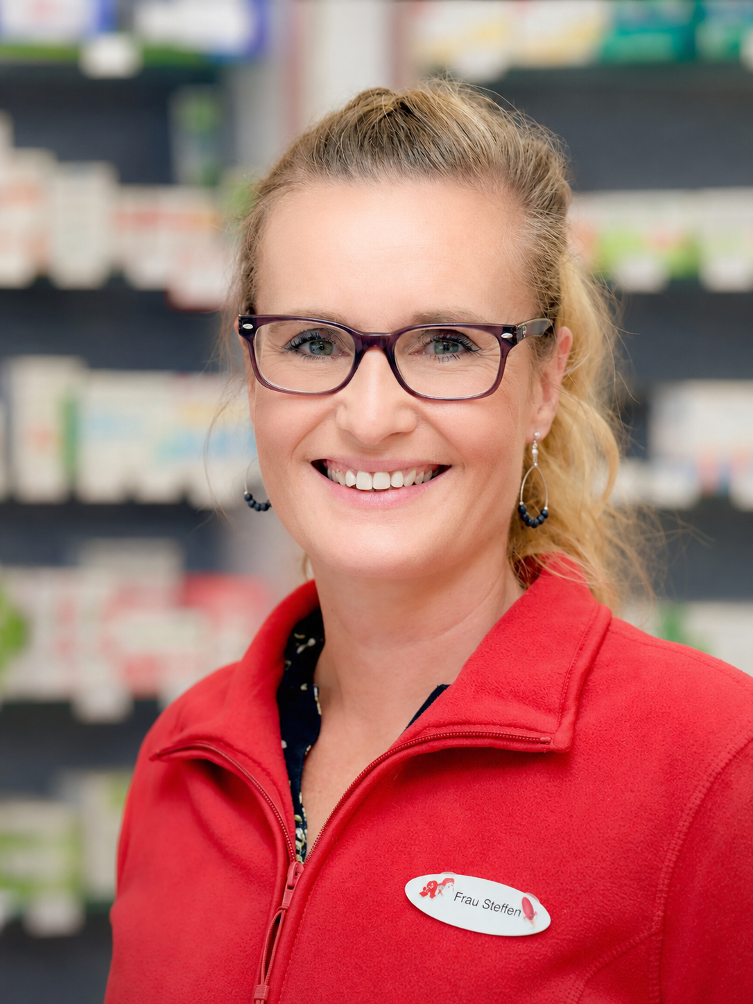 Frau in rotem Hemd, Brille, lächelnd, in einer Apotheke.