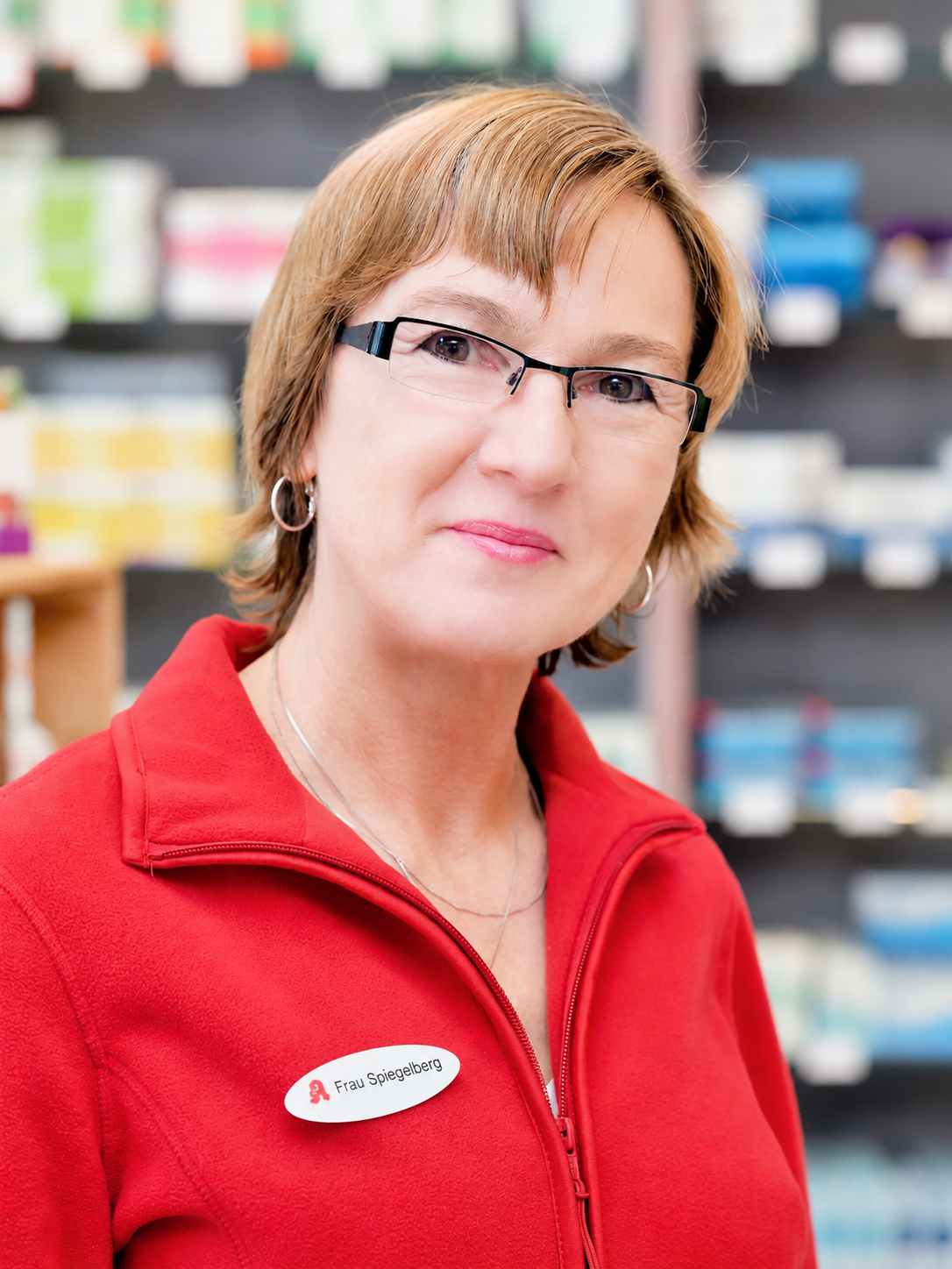 Frau in rotem Hemd und Brille lächelt in einer Apotheke und trägt ein Namensschild.