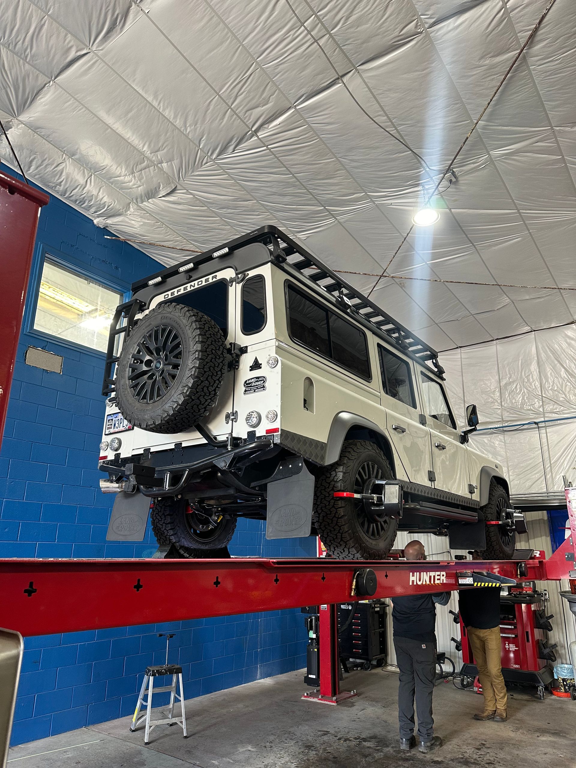 White Land Rover Defender on a lift in a garage, two people working, silver ceiling | Marshside Motors