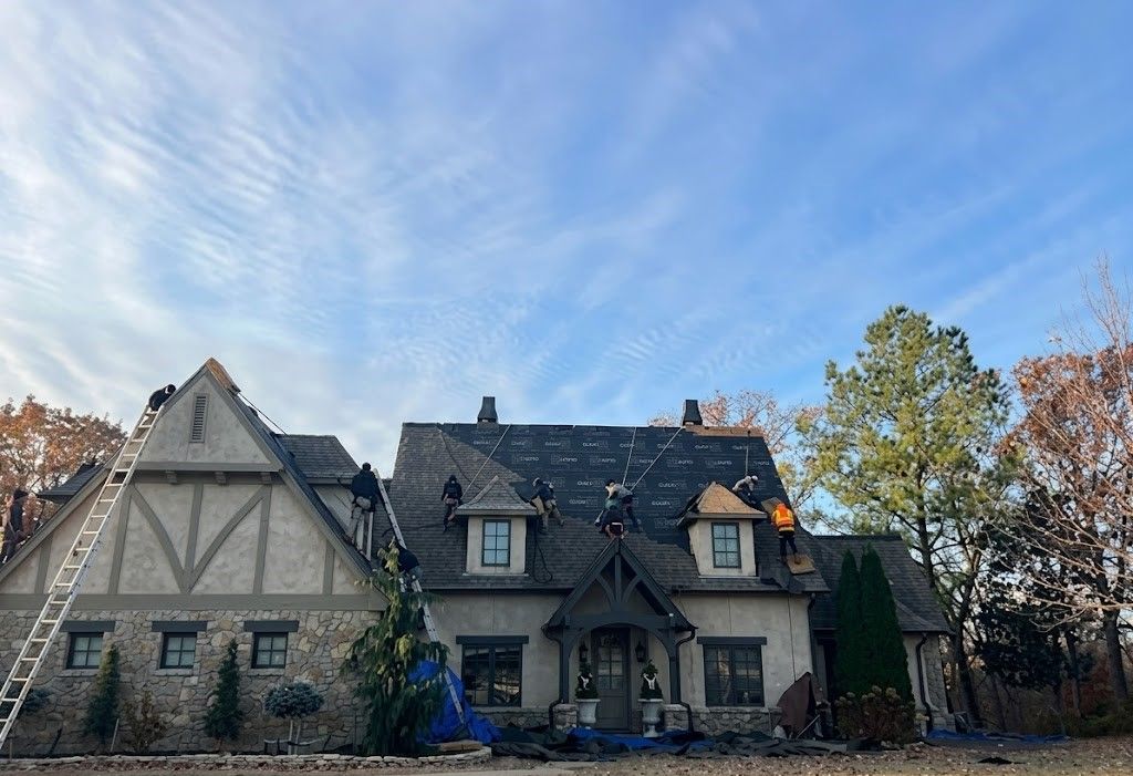 Two-story stone-and-timber house with dark roof, set under a bright blue sky with wispy clouds.