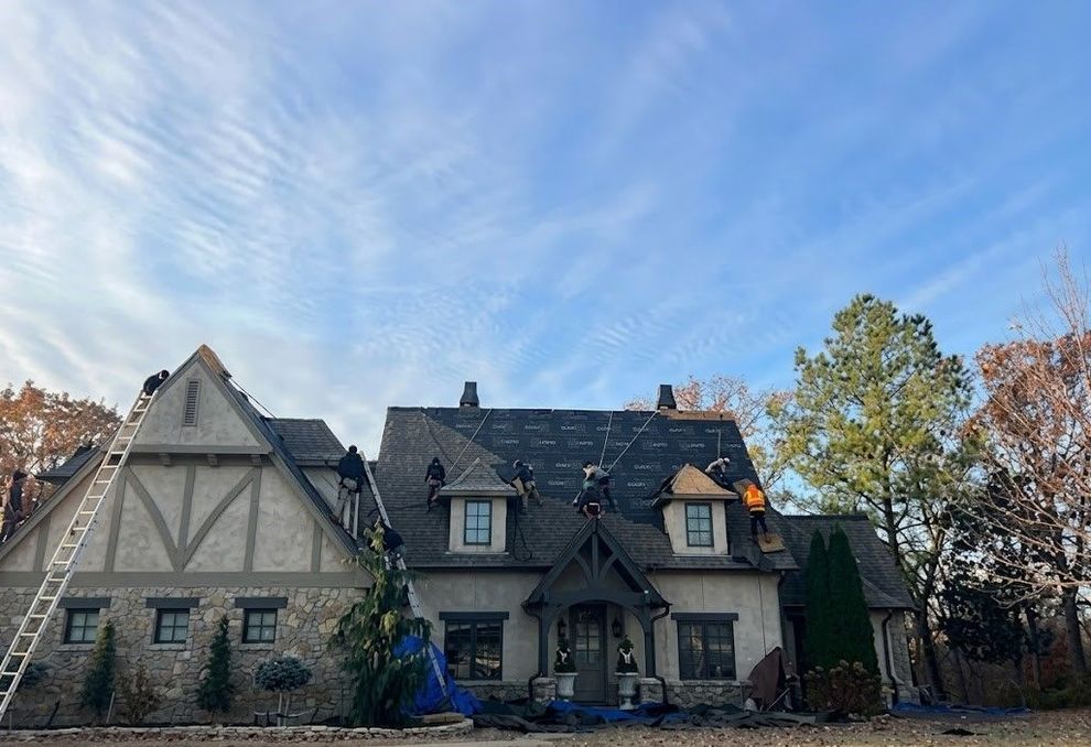 Two-story stone house with steep roofs and dormers beneath a blue sky, framed by trees and bare branches