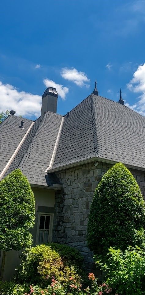 Stone house with steep gray roof and manicured green shrubs under a bright blue sky