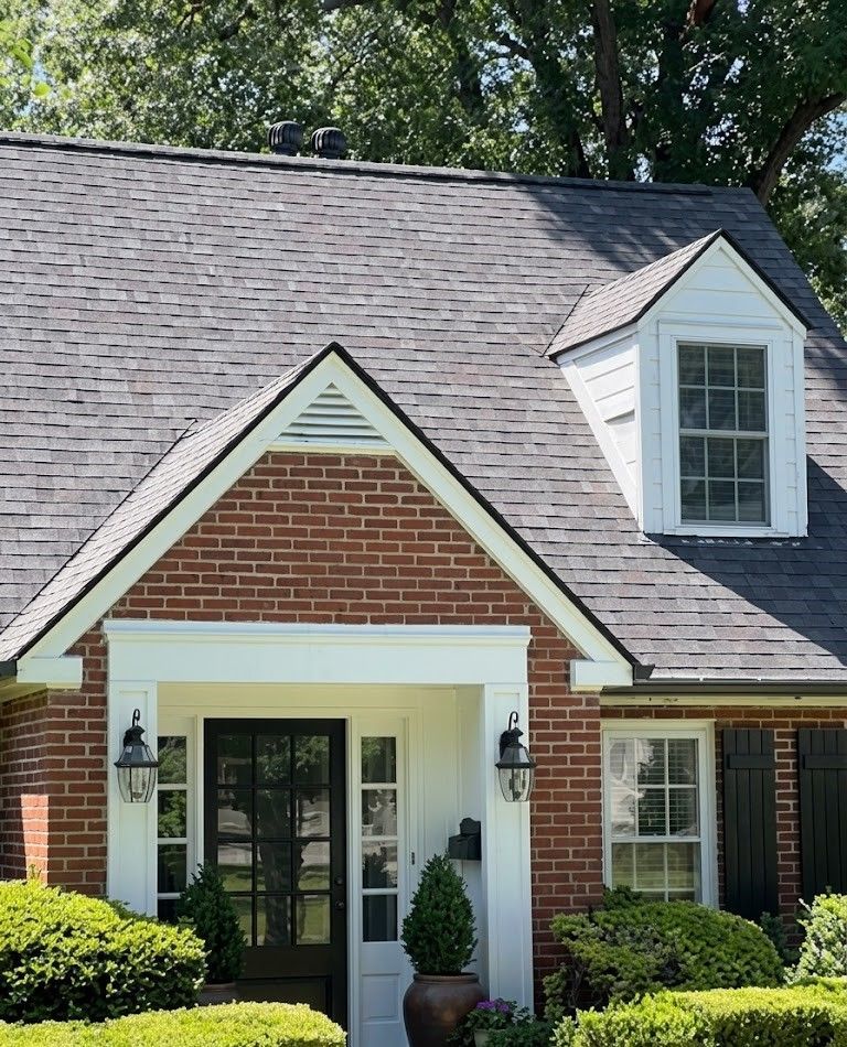 Brick house front entrance with white trim, black door, and dormer window beneath a shingled roof