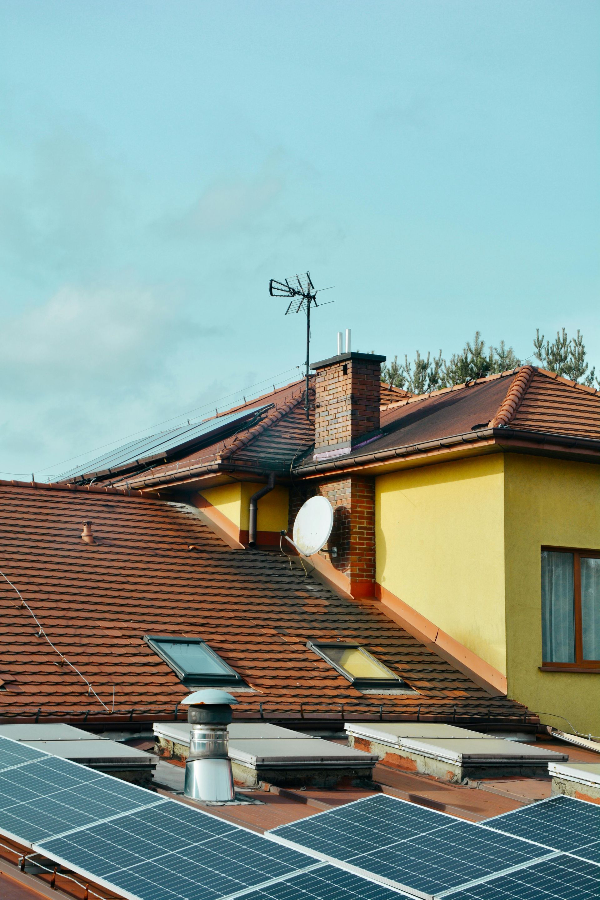 Solar panels on a red-tiled rooftop with a chimney and antenna under a blue sky.