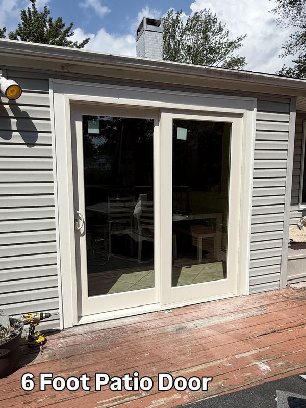 A new 6-foot cream-colored sliding patio door installed in a house with light gray vinyl siding and a wood deck.