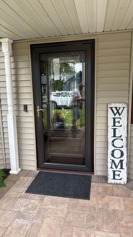 A brown storm door on a house with beige siding, a grey welcome mat, and a vertical 