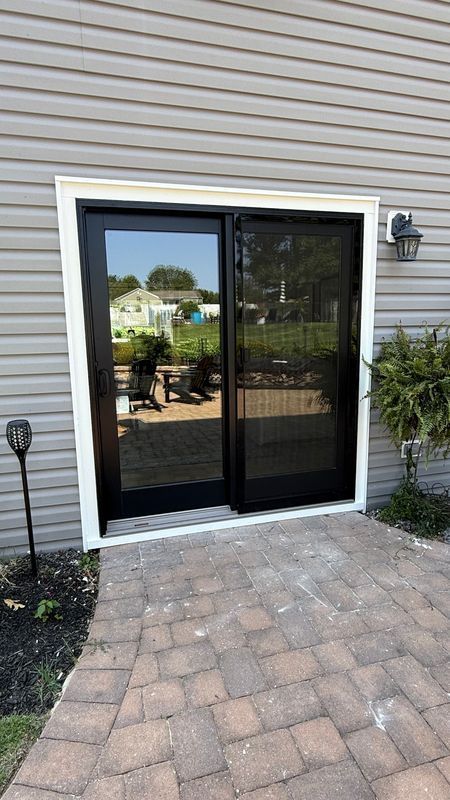 A black-framed sliding glass patio door set in beige vinyl siding above a paved stone walkway.