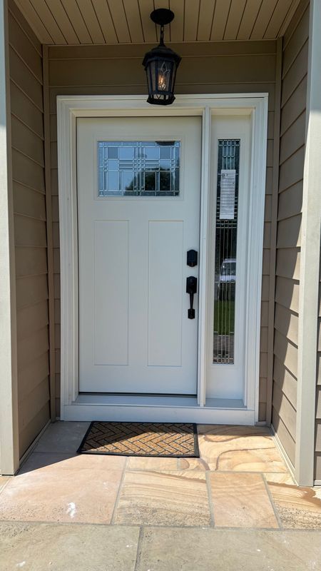 A white front door with a window and sidelight, mounted in tan siding above a stone patio with a doormat.