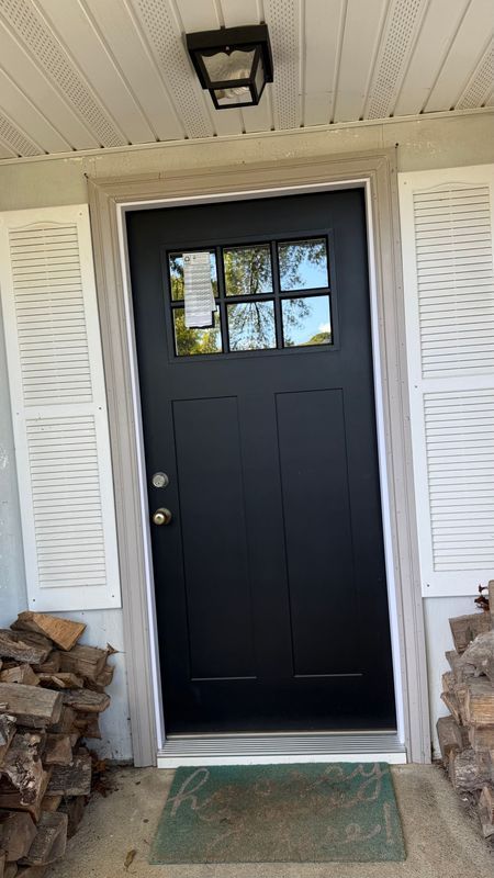 A black front door with a six-pane window, flanked by white shutters and stacks of firewood on a covered porch.