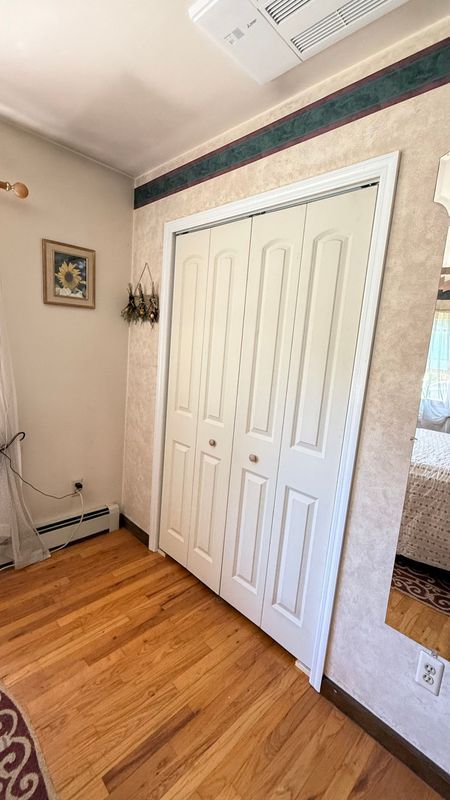 White bi-fold closet doors in a room with wood floors, light patterned wallpaper, and a framed sunflower picture.