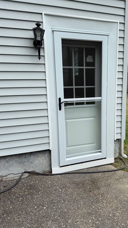 A white storm door with a window and black handle installed on a house with light grey vinyl siding and a concrete porch.