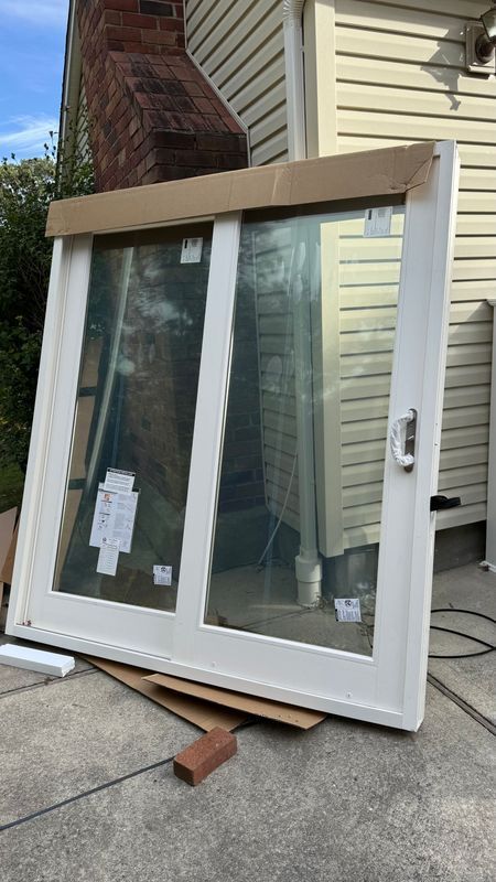 A new white sliding glass door unit leaning against the side of a house with beige siding and a brick chimney.