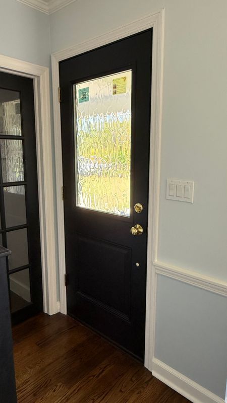 A black front door with a glass pane, brass hardware, and white trim in a light blue room with dark wood flooring.