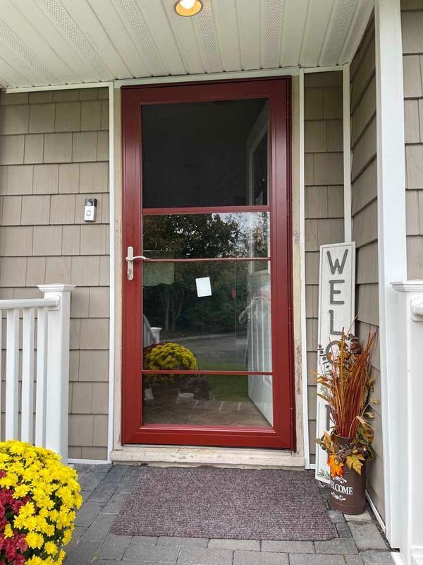 A red storm door with glass panels on a porch, framed by tan shingle siding, a welcome sign, and potted mums.