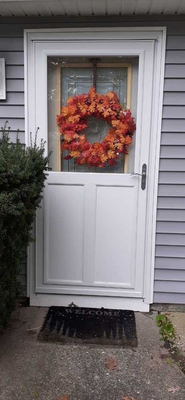 A white storm door with an autumn-colored wreath, positioned on a house with gray siding and a welcome mat.