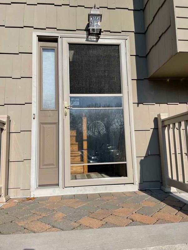 A brown front door with a glass storm door and adjacent sidelight, set in a tan-sided home with a paved porch.