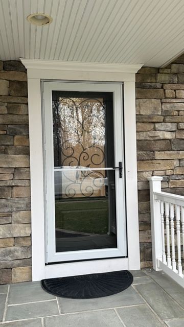 A white storm door with a scrollwork design on the glass, set in a stone-walled porch entrance with a black half-moon mat.