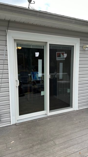 A white-framed sliding glass door installed on the exterior of a home with light gray siding and a wooden deck.