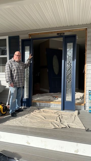 A person in a plaid shirt stands on a porch next to a blue door frame being installed in an open house entryway.