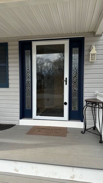 A white storm door with glass panels is set within a dark blue frame on a gray porch with light gray siding.