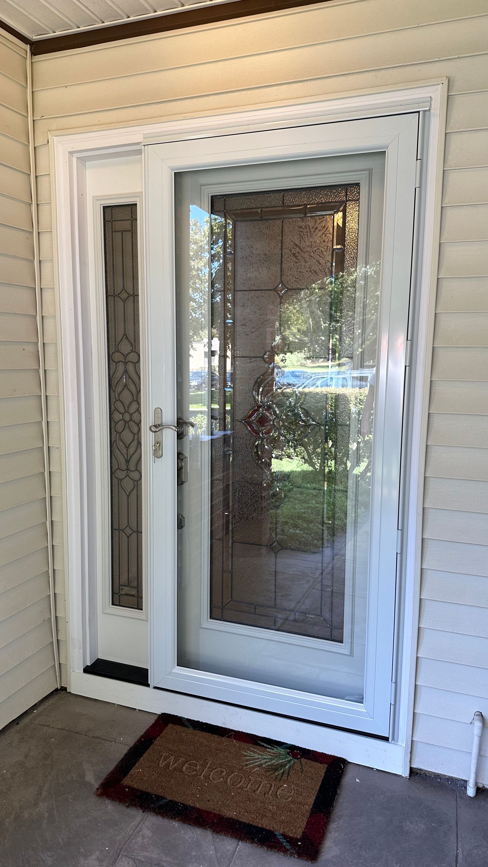 White front storm door with a decorative glass panel and a side light, mounted on a porch with beige horizontal siding.