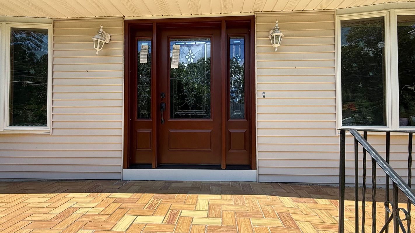 A brown front door with glass panels and sidelights on a house with light-colored siding and a brick-patterned porch.