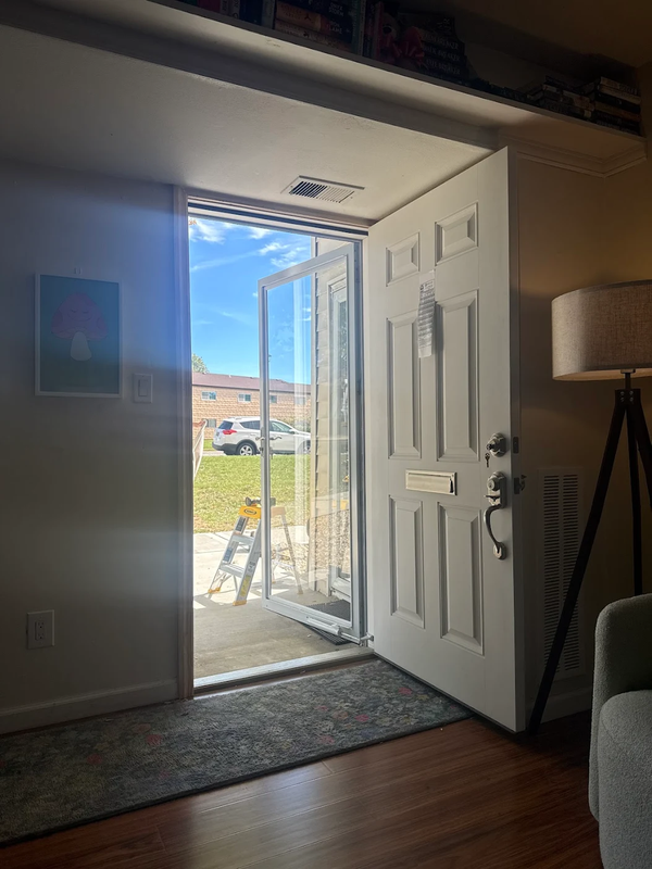 An open white front door reveals a sunny residential exterior with a lawn, a parked car, and a step ladder.
