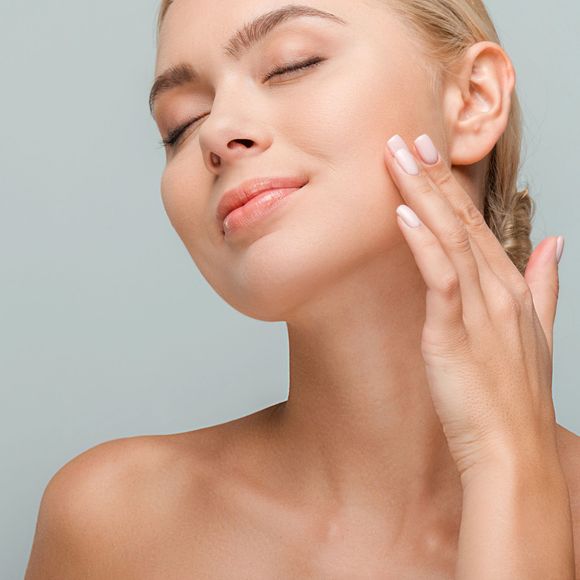 Donna con gli occhi chiusi che si applica una crema per la cura della pelle sulla guancia, su uno sfondo grigio chiaro.