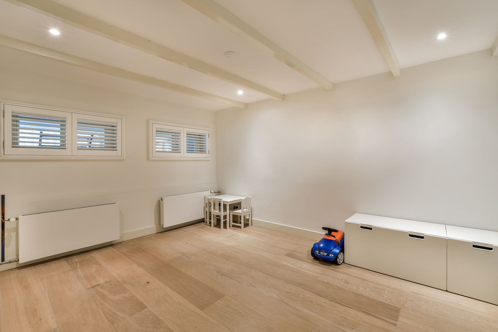 Empty playroom with light wood floors, white walls and ceiling with exposed beams, small table and chairs, toy car.