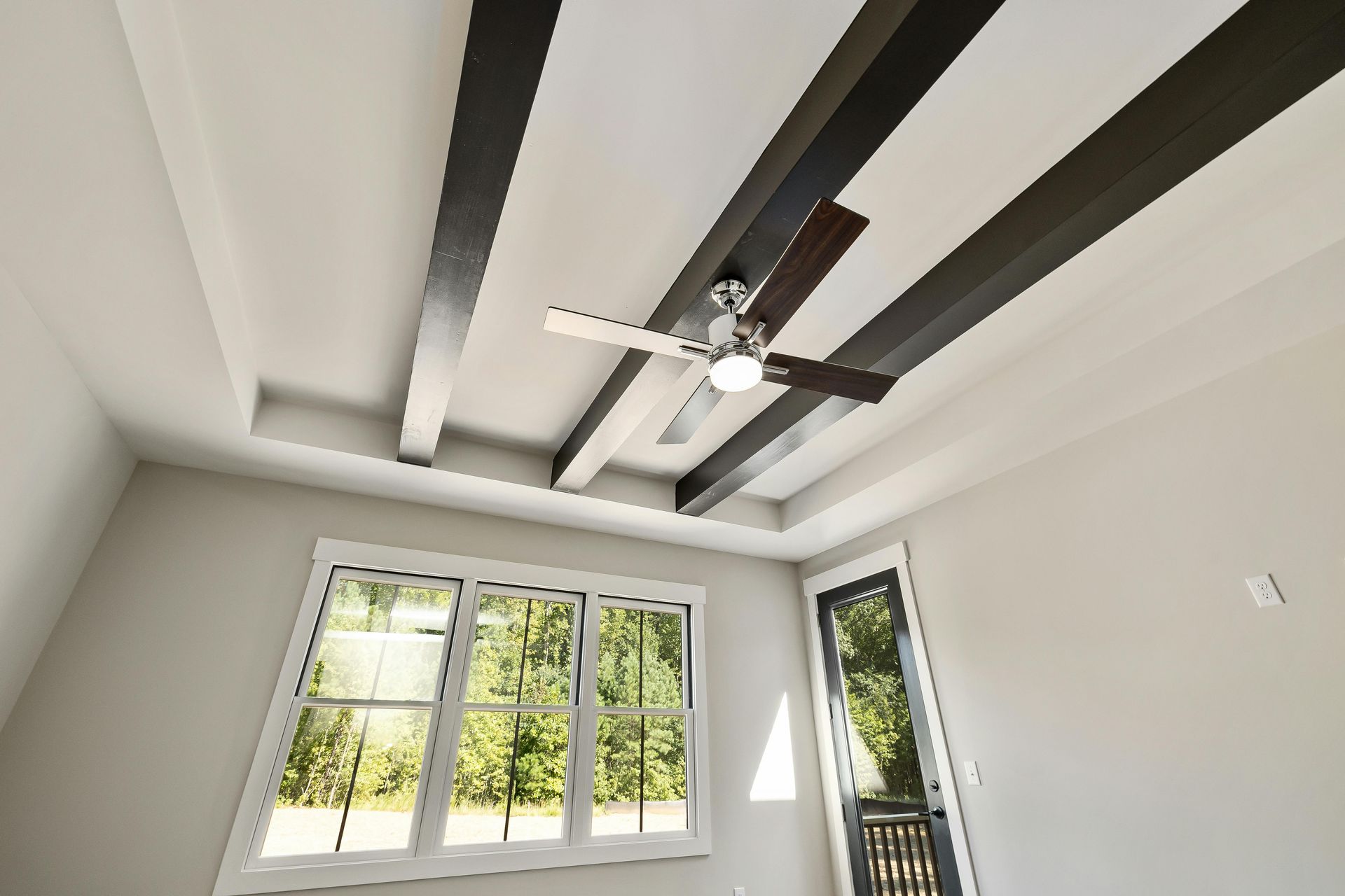 Room with white walls, a ceiling fan, and dark beams. A window overlooks trees.
