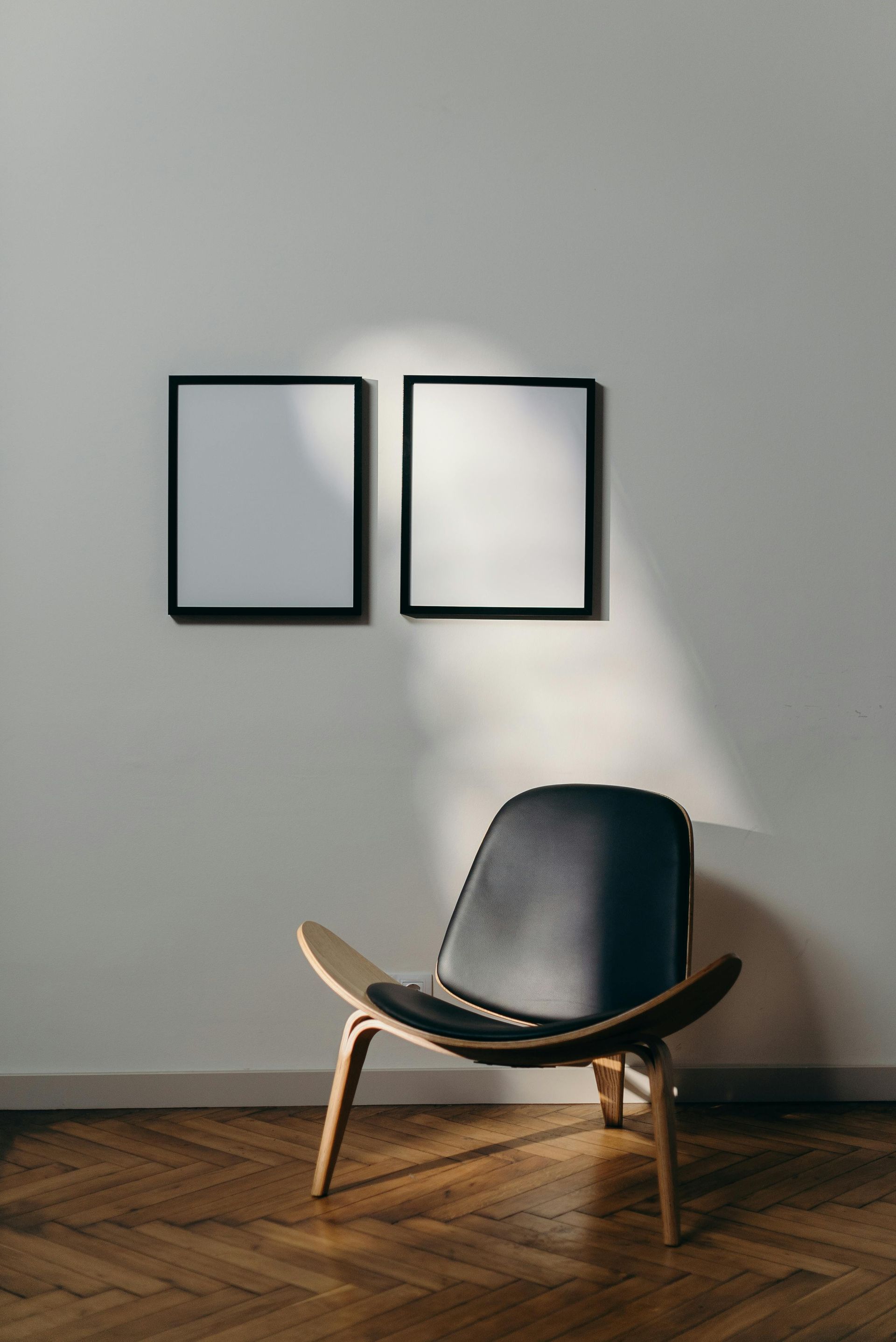 A black-framed chair sits beneath two framed white pictures on a white wall; sunlit hardwood floors.