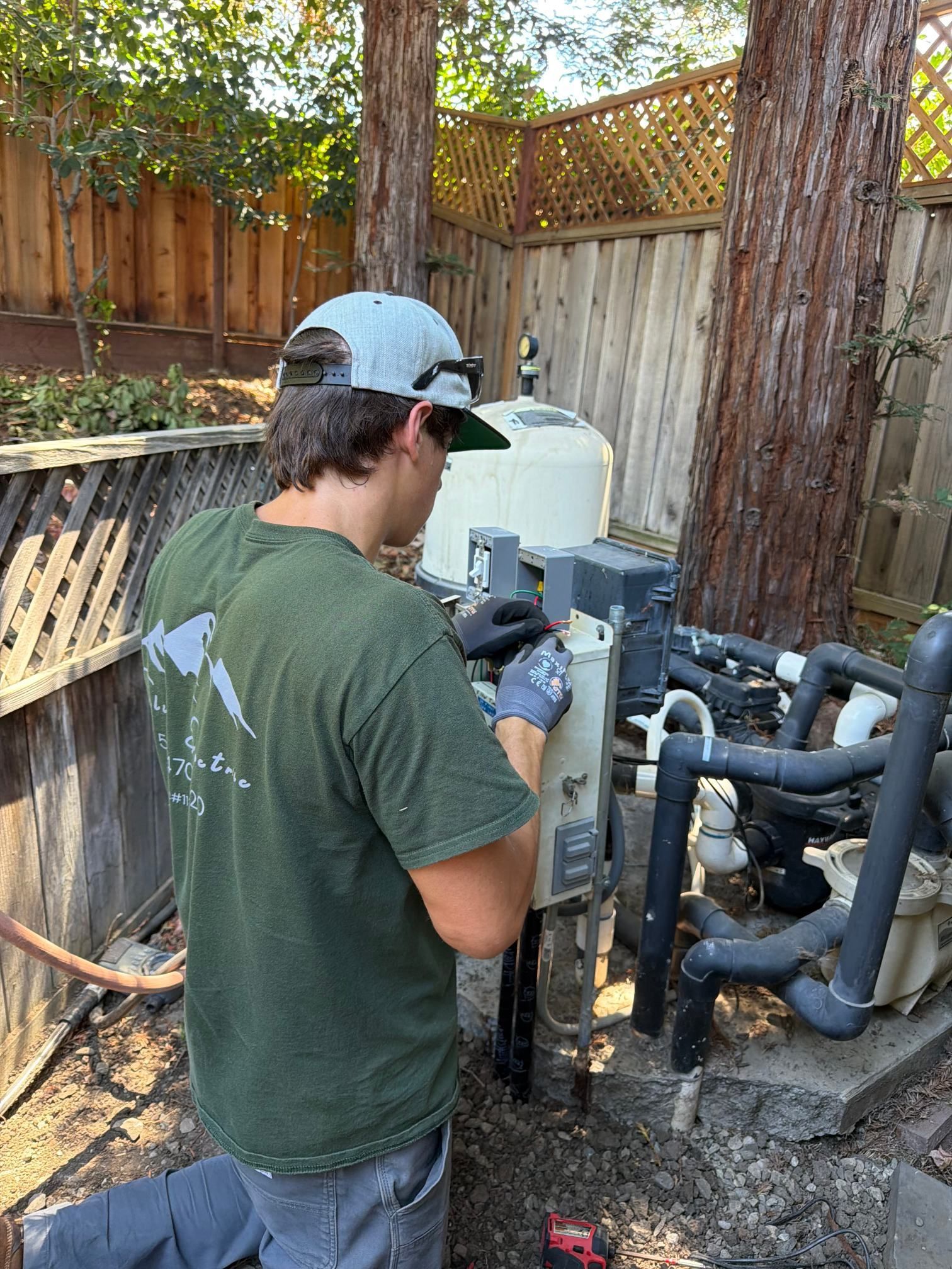 Worker inspecting pool equipment outdoors beside a wooden fence and trees