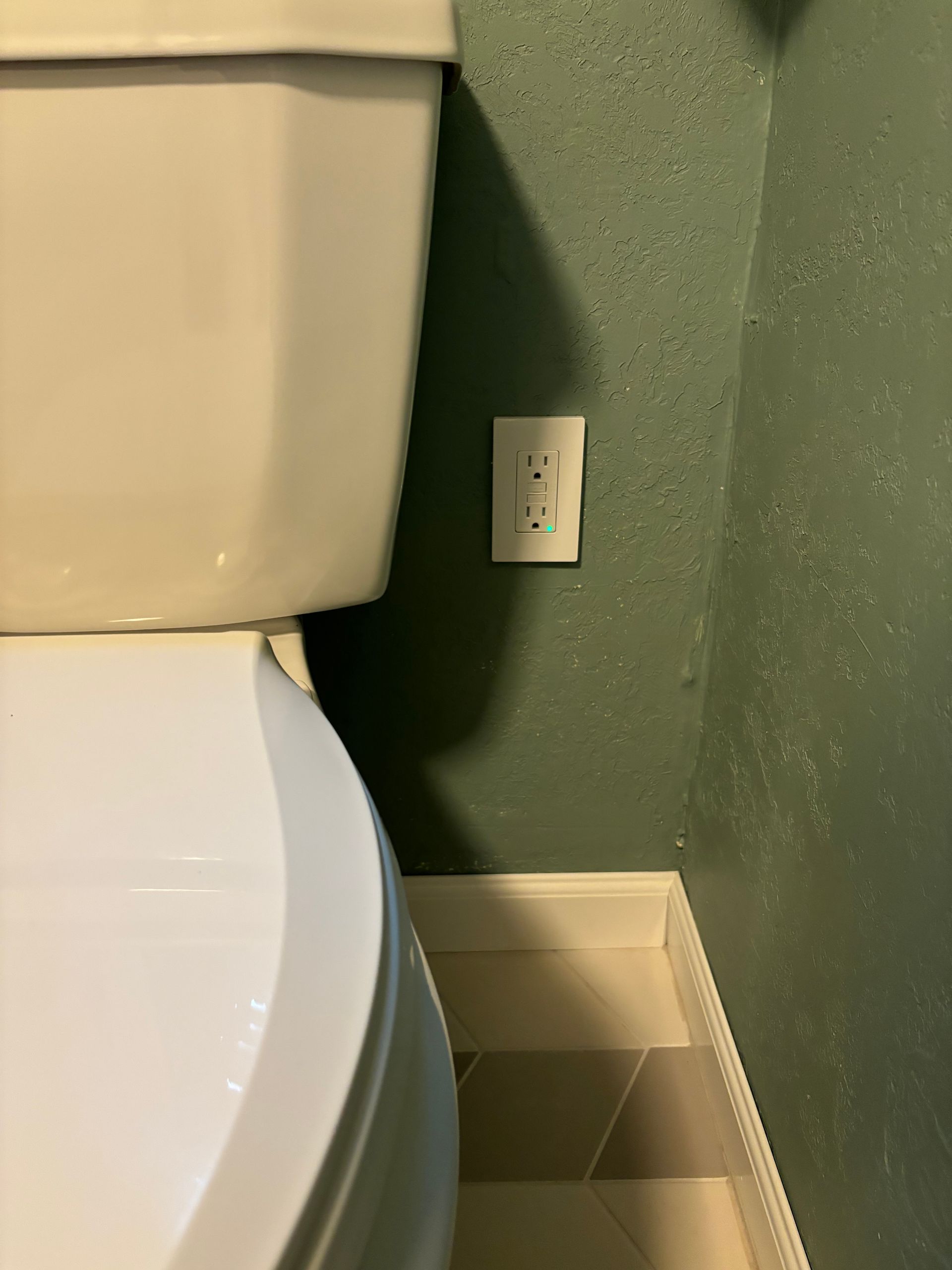 Toilet next to a green wall with an electrical outlet near the floor.
