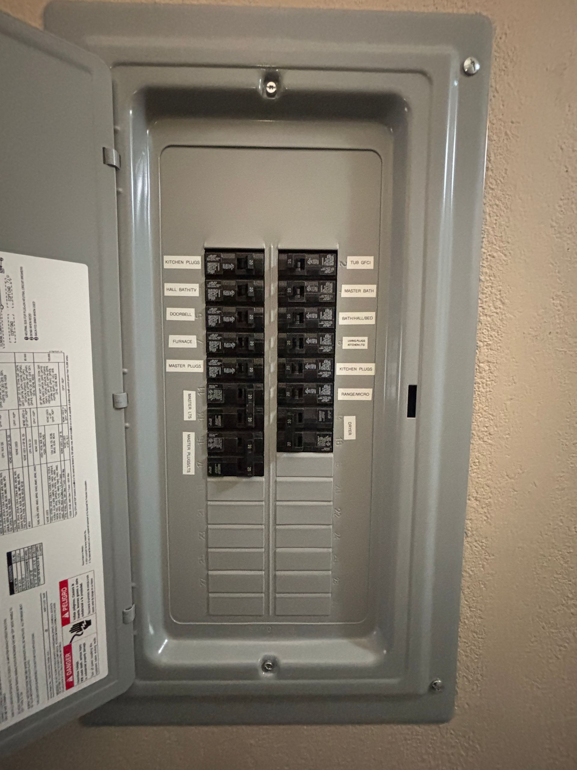 Open electrical panel with circuit breakers. Beige wall, gray metal panel. Breakers are labeled.
