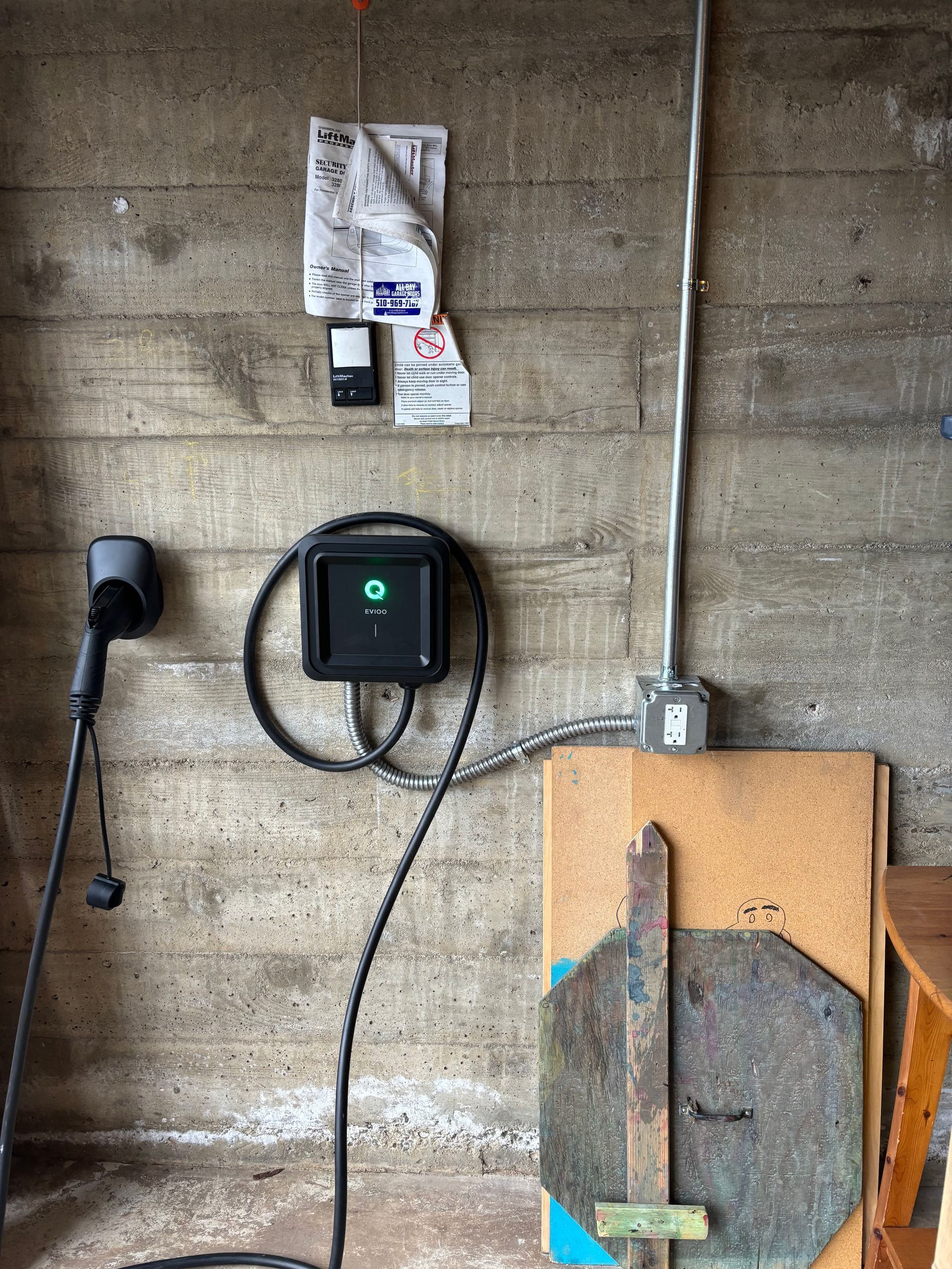 Black electric vehicle charger on concrete wall with attached cord and documents.