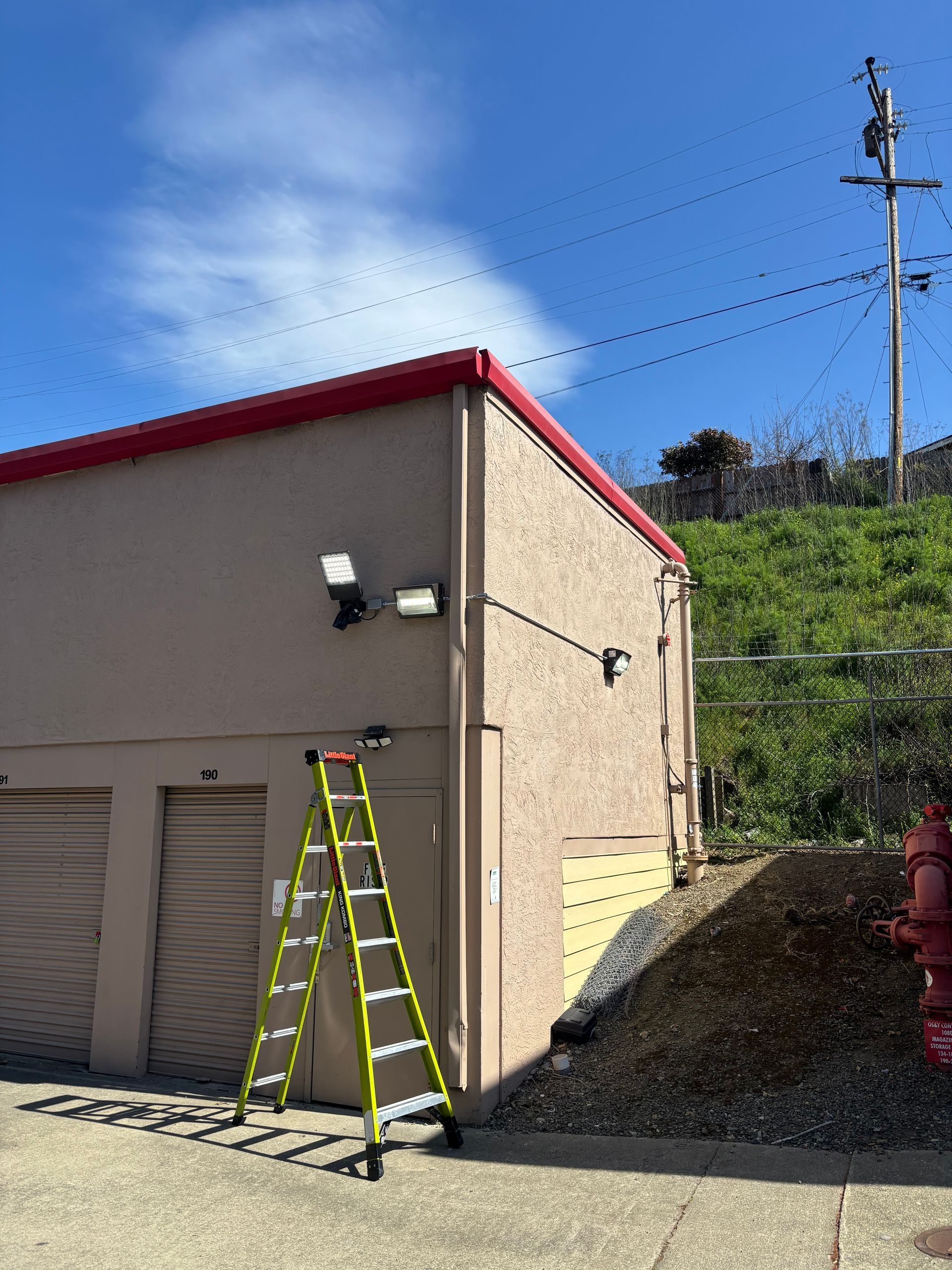 Exterior of a beige storage building with a ladder, overhead lights, and a utility pole against a blue sky.