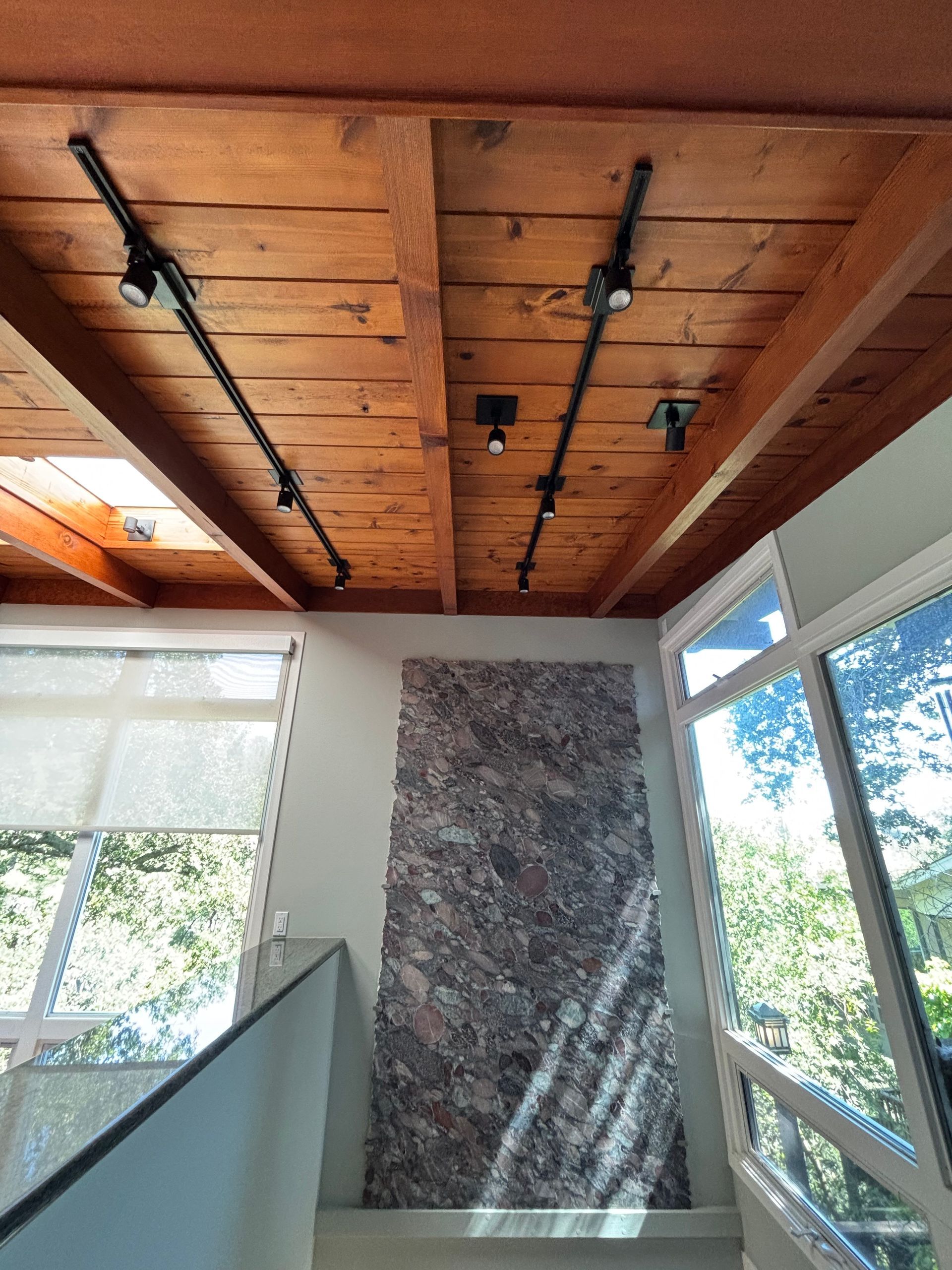 Wooden beam ceiling with track lighting, above a stairway with a textured wall and windows.