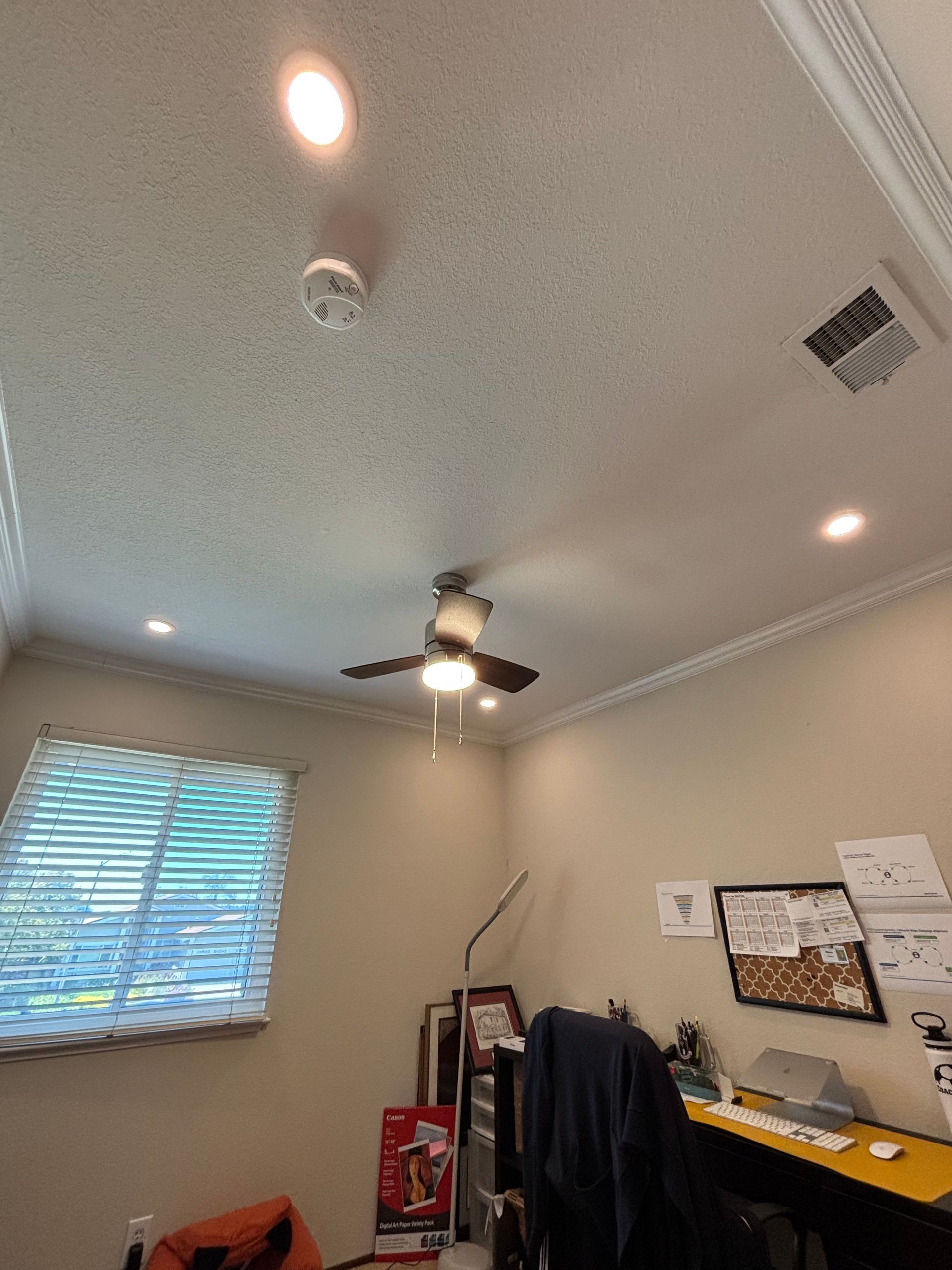 Ceiling with fan, lights, smoke detector, and vent. Window and desk visible in a room with tan walls.
