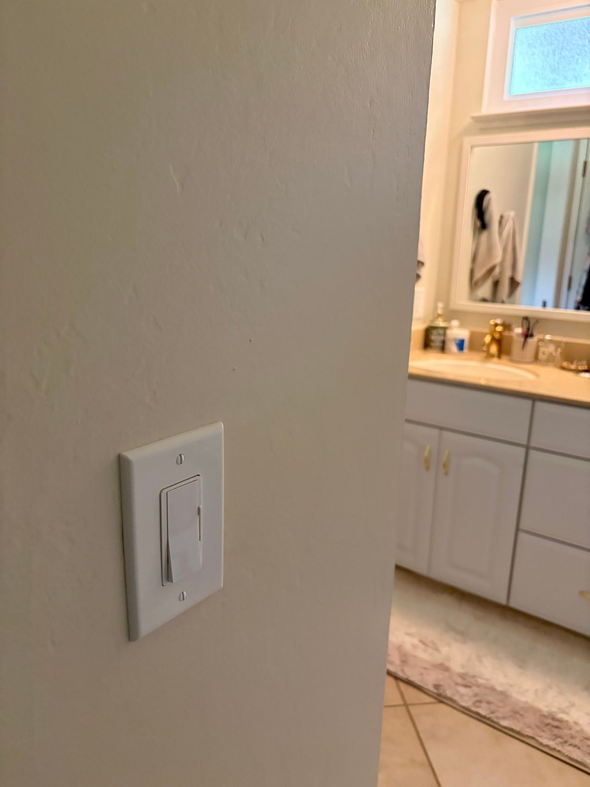 Wall-mounted light switch beside a bathroom vanity and mirror, with beige walls and white cabinets.