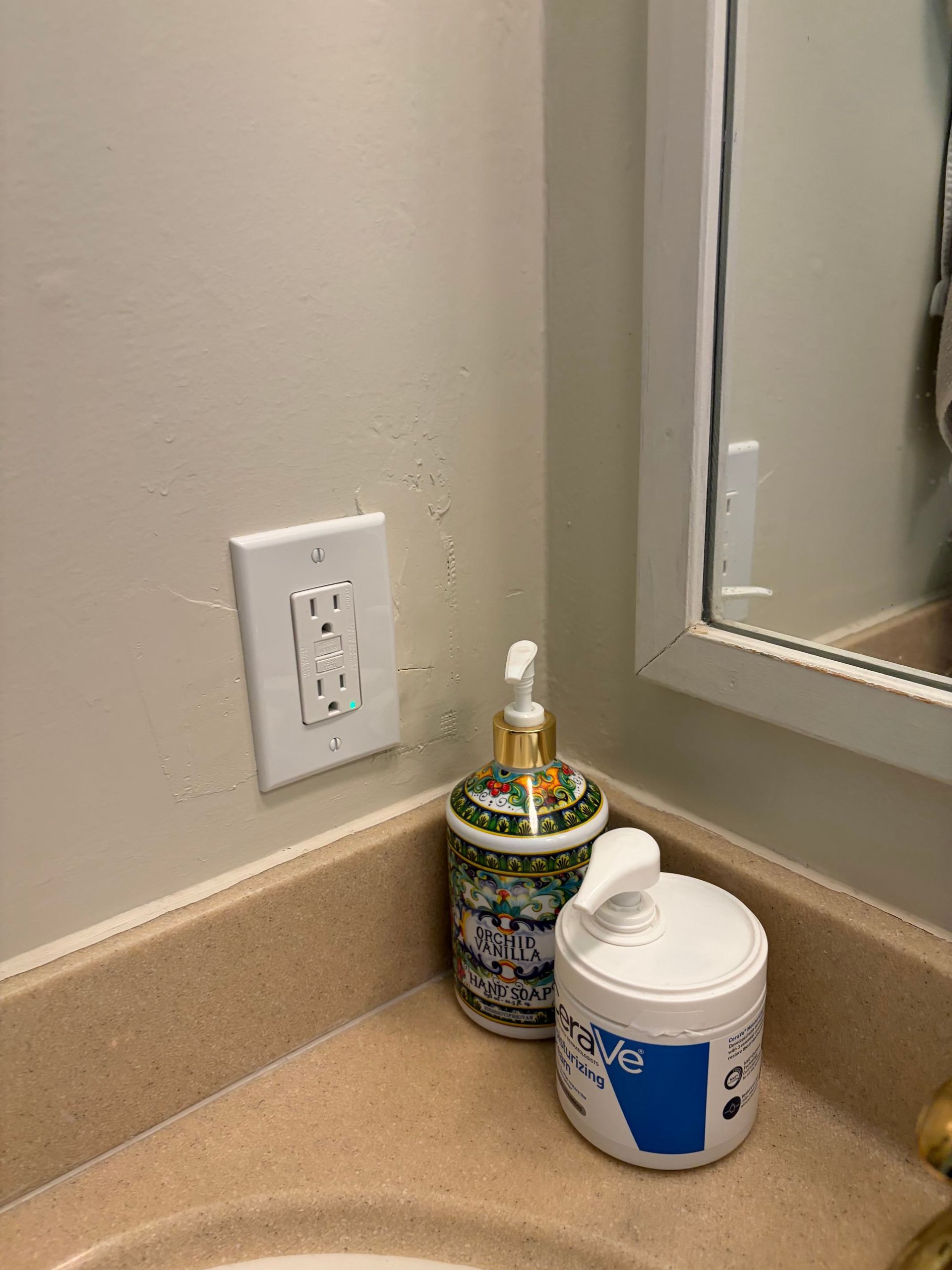 Bathroom vanity corner with outlet, mirror edge, lotion pump, and a white jar on a beige countertop